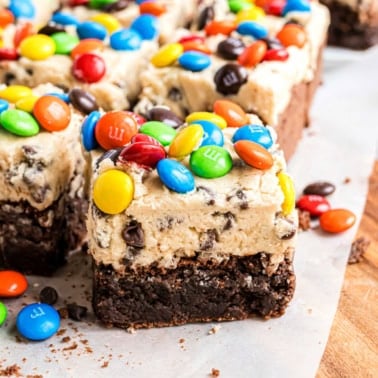 Brownie bars with a cookie dough layer, topped with colorful M&M candies on parchment paper.