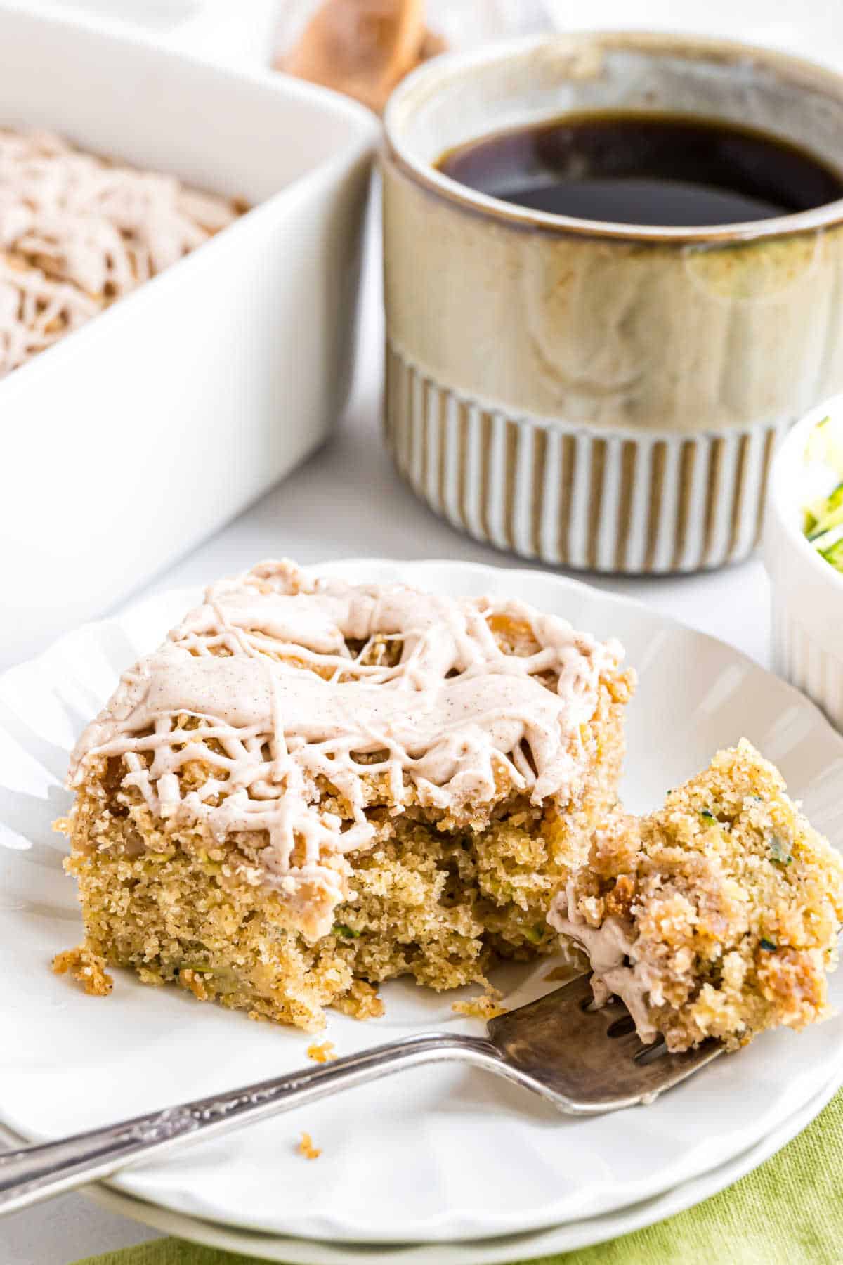 A slice of frosted cake on a plate with a fork, next to a mug of coffee.