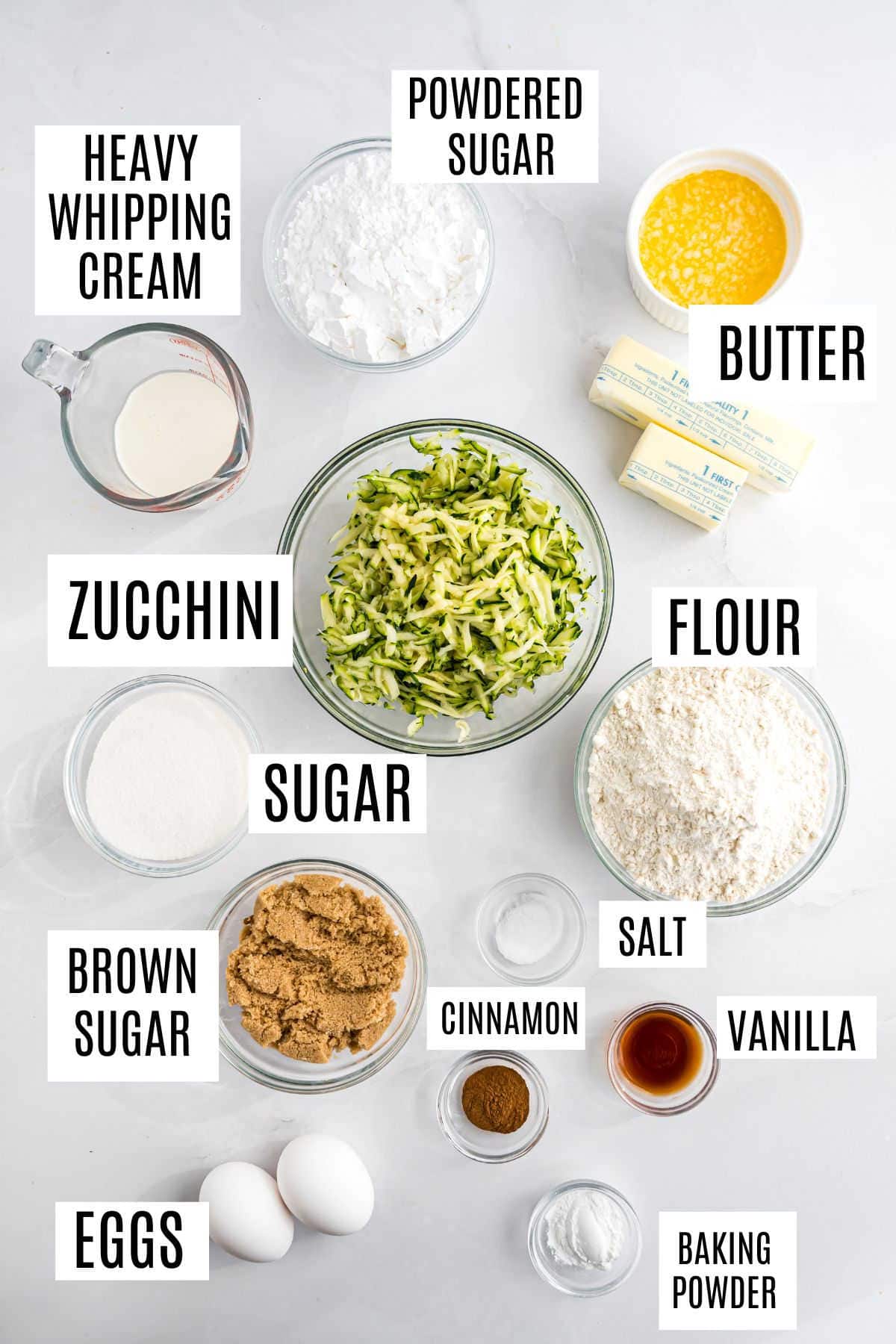 Labeled baking ingredients including zucchini, flour, sugar, eggs, spices, and butter on a white surface.