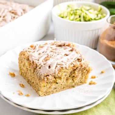 A slice of crumb-topped cake with icing on a white plate, with shredded zucchini and cinnamon nearby.