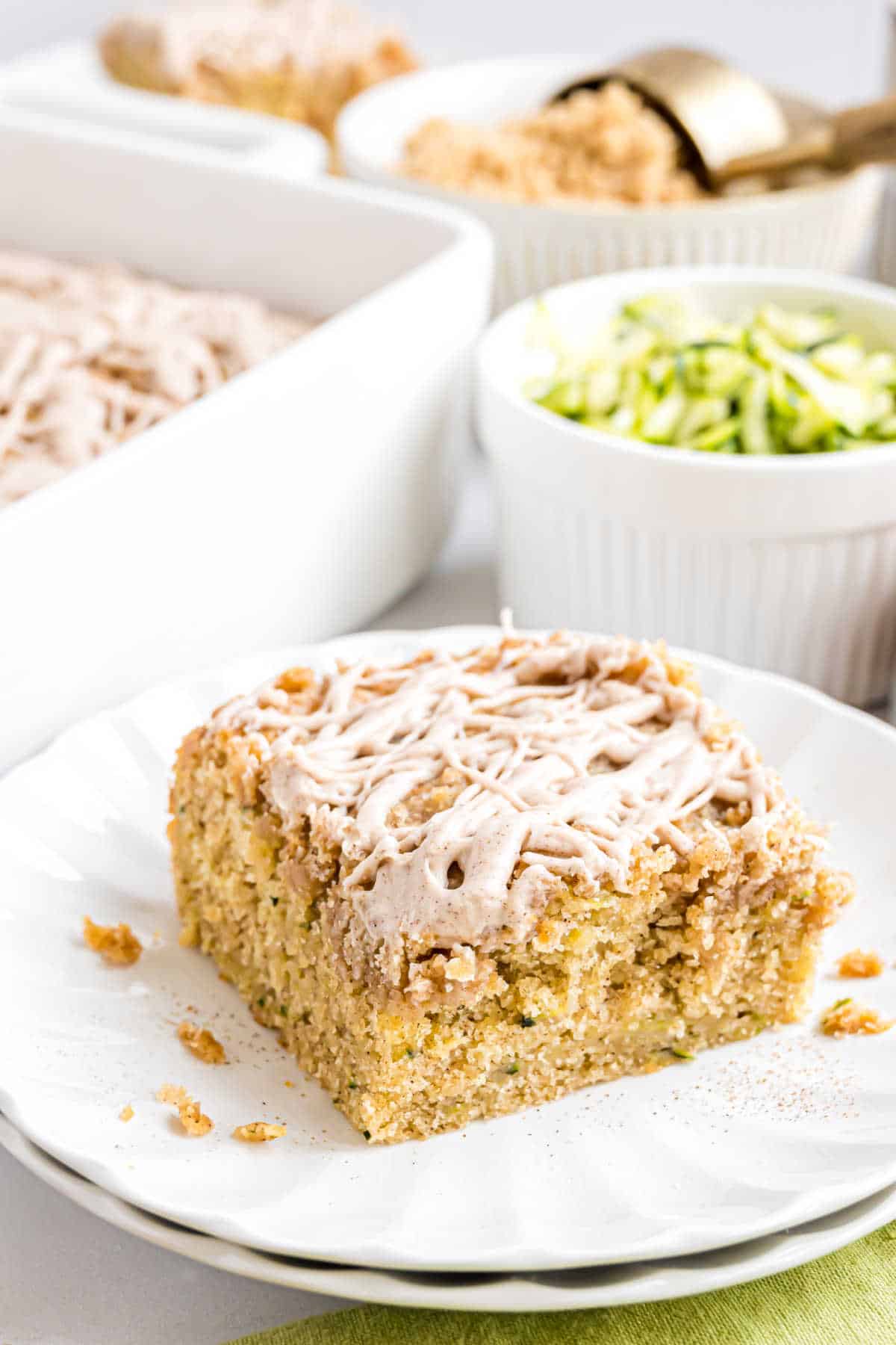 A slice of crumb-topped zucchini cake with icing on a white plate, surrounded by baking ingredients.