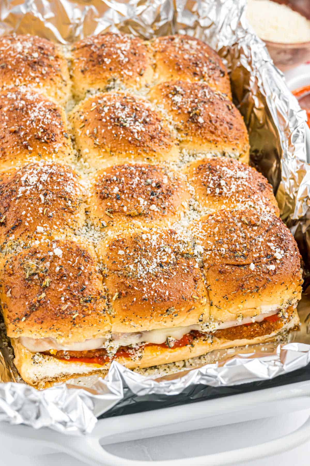 A pan of baked Italian sliders with melted cheese and herbs, sliced into squares on foil.