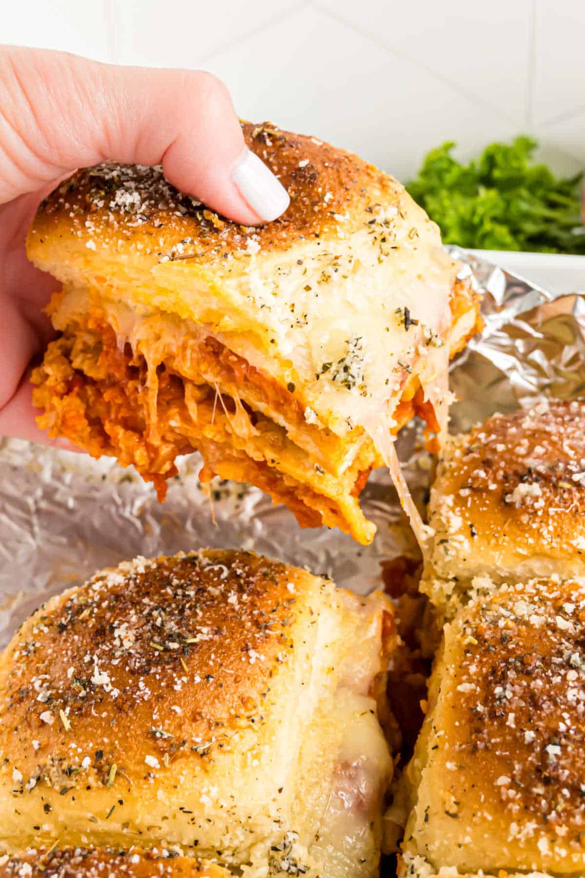 A hand holding a cheesy chicken parmesan slider with golden, seasoned buns and melted cheese.