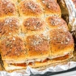 A tray of baked slider sandwiches with seasoned tops, filled with cheese and sauce, on foil.