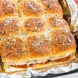 A tray of baked slider sandwiches with seasoned tops, filled with cheese and sauce, on foil.