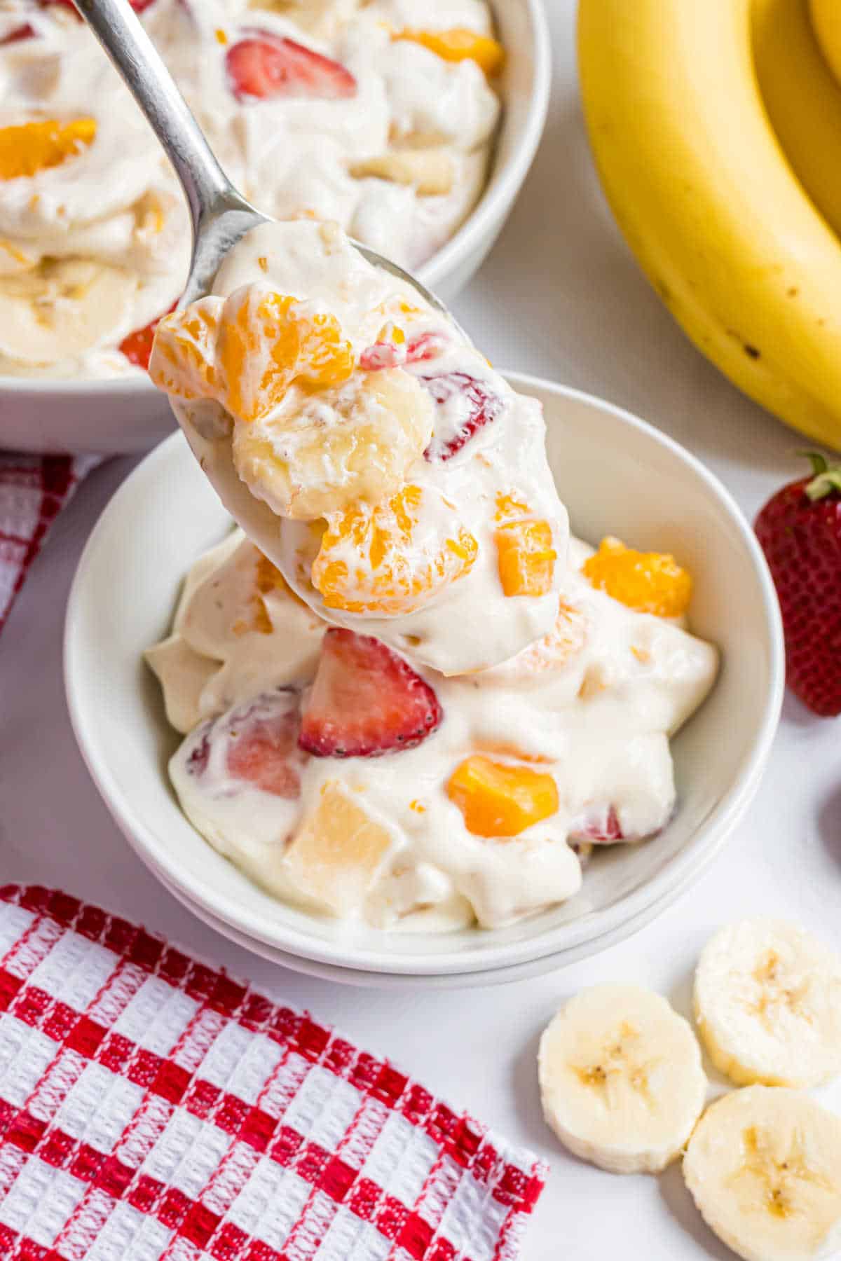 A bowl of creamy fruit salad with bananas, strawberries, and oranges, with a spoon lifting a serving.