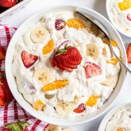 A bowl of creamy fruit salad with bananas, strawberries, and orange slices, garnished with a whole strawberry.