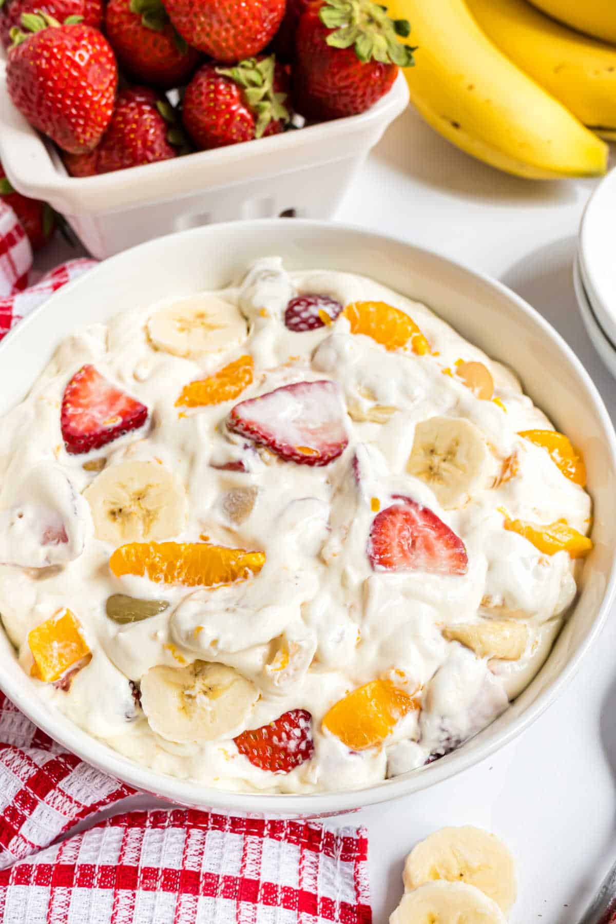A bowl of creamy fruit salad with bananas, strawberries, and oranges, surrounded by fresh fruit.