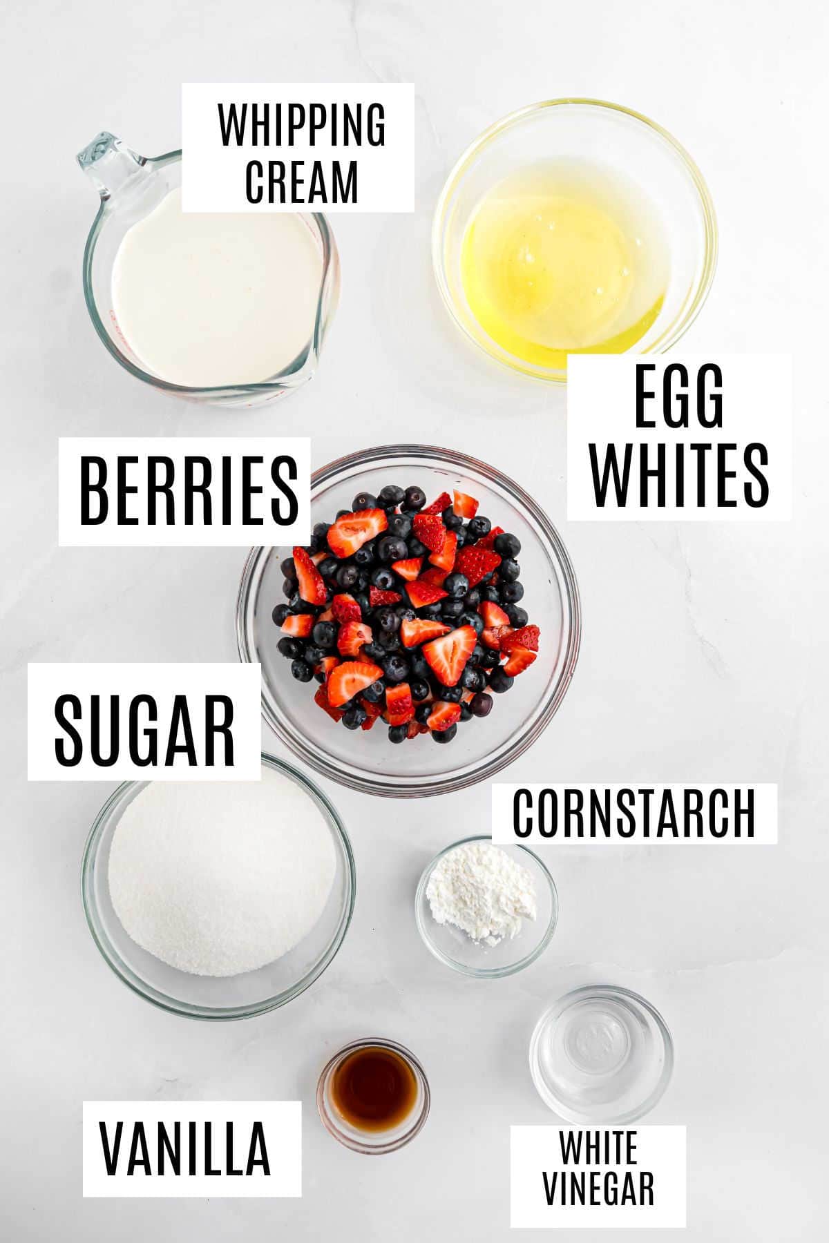 Bowls of whipping cream, egg whites, mixed berries, sugar, cornstarch, vanilla, and white vinegar on a white surface.