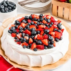 A pavlova topped with whipped cream, fresh blueberries, and chopped strawberries on a parchment-lined plate.