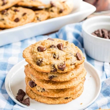 A stack of chocolate chip cookies on a plate, with more cookies and chocolate chips in the background.