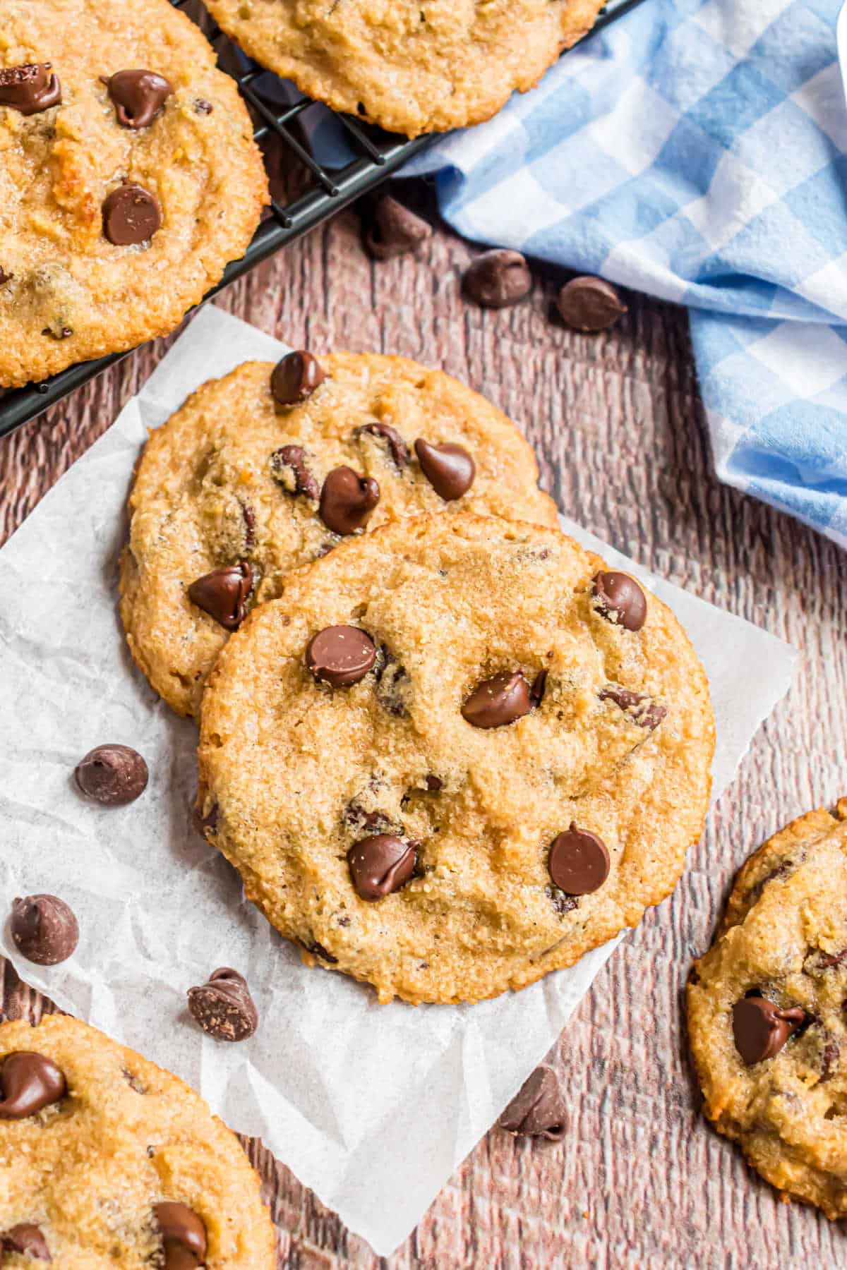 Chocolate chip cookies on parchment paper with scattered chocolate chips and a checkered cloth nearby.