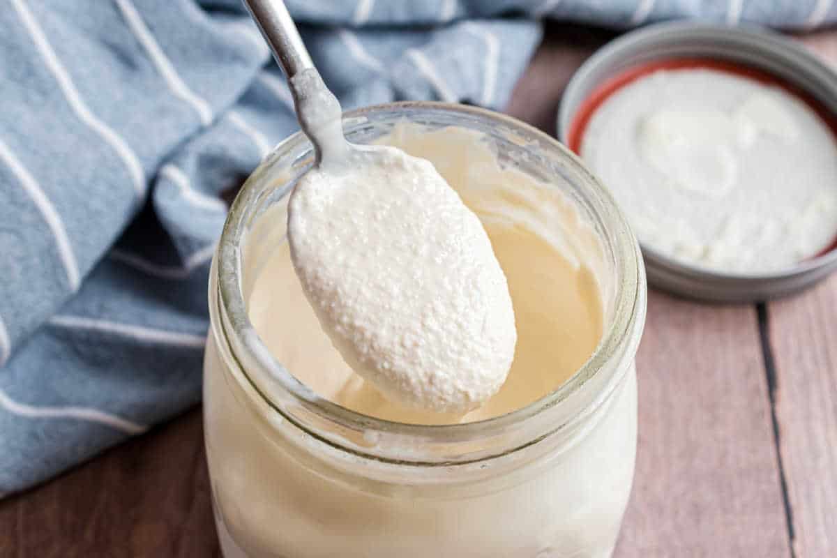 A spoonful of creamy yogurt held over an open glass jar on a wooden surface.