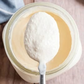 A spoonful of thick, creamy yogurt held over an open jar on a wooden surface.