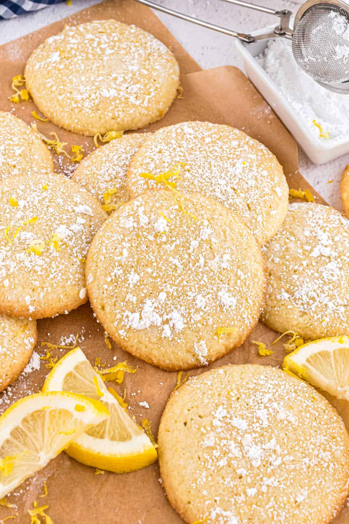 Round lemon cookies dusted with powdered sugar, surrounded by lemon slices and zest on brown parchment paper.