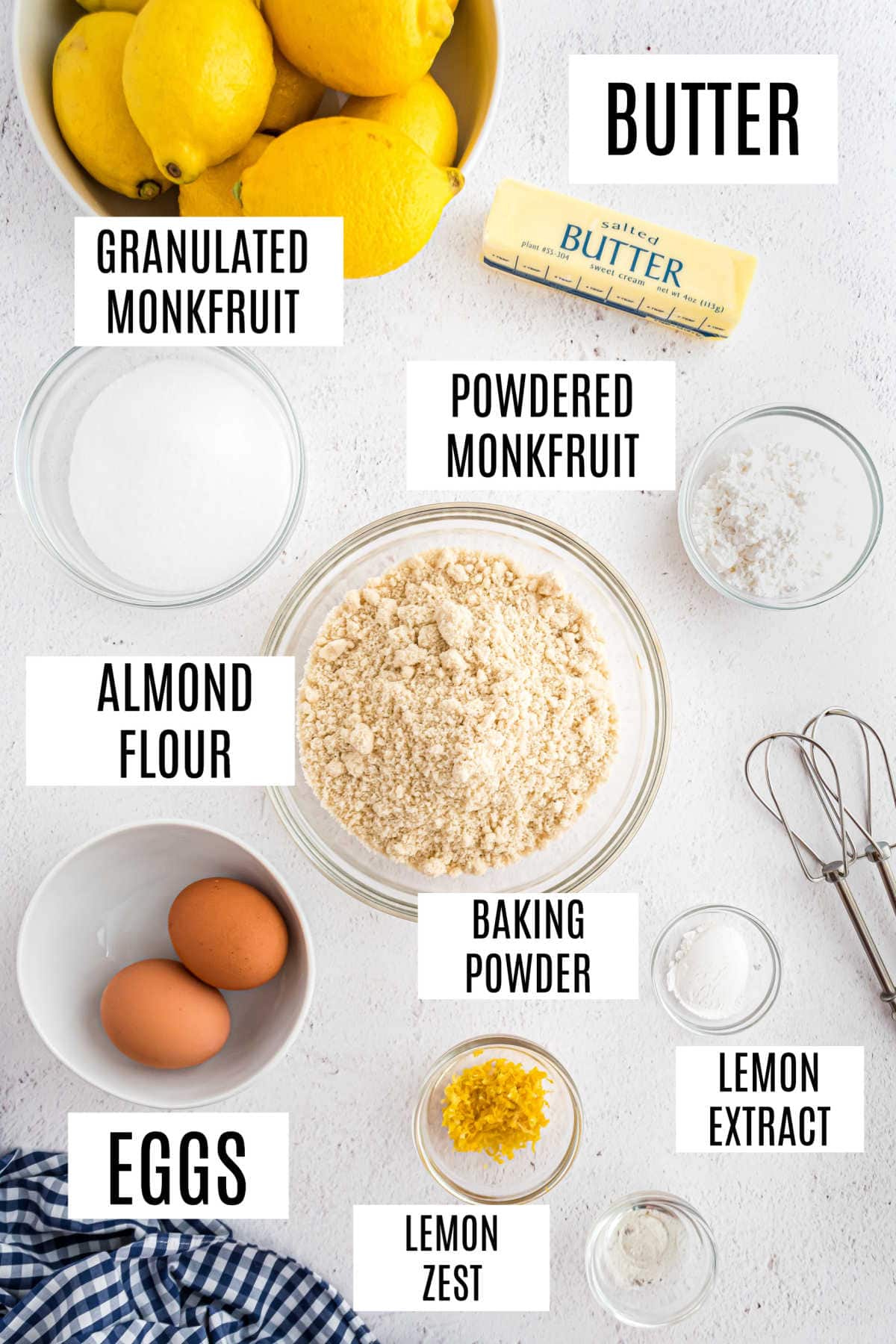 Labeled baking ingredients on a white surface: lemons, butter, almond flour, eggs, sweeteners, and baking powder.
