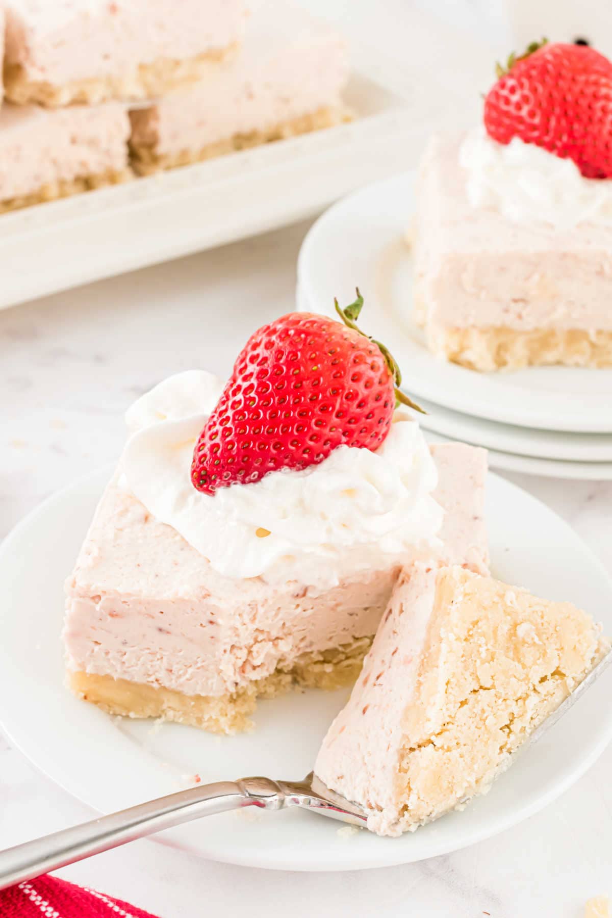 A strawberry cheesecake bar topped with whipped cream and a fresh strawberry, with a forkful on a white plate.