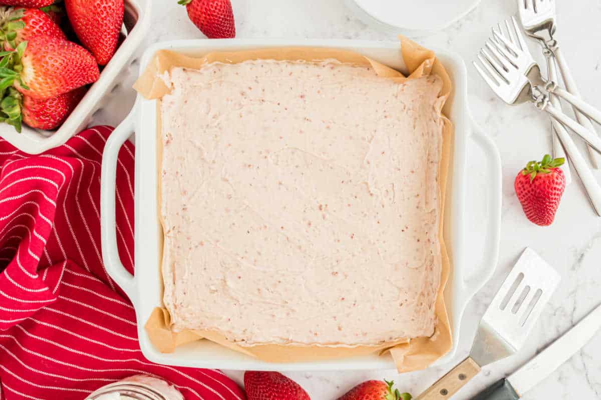 Square baking dish with strawberry mixture, surrounded by fresh strawberries and kitchen utensils.