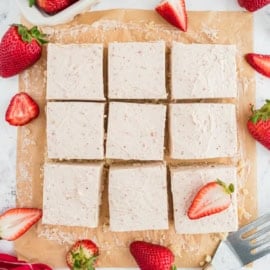 Nine frosted strawberry bars on parchment paper, surrounded by fresh strawberries, some sliced.