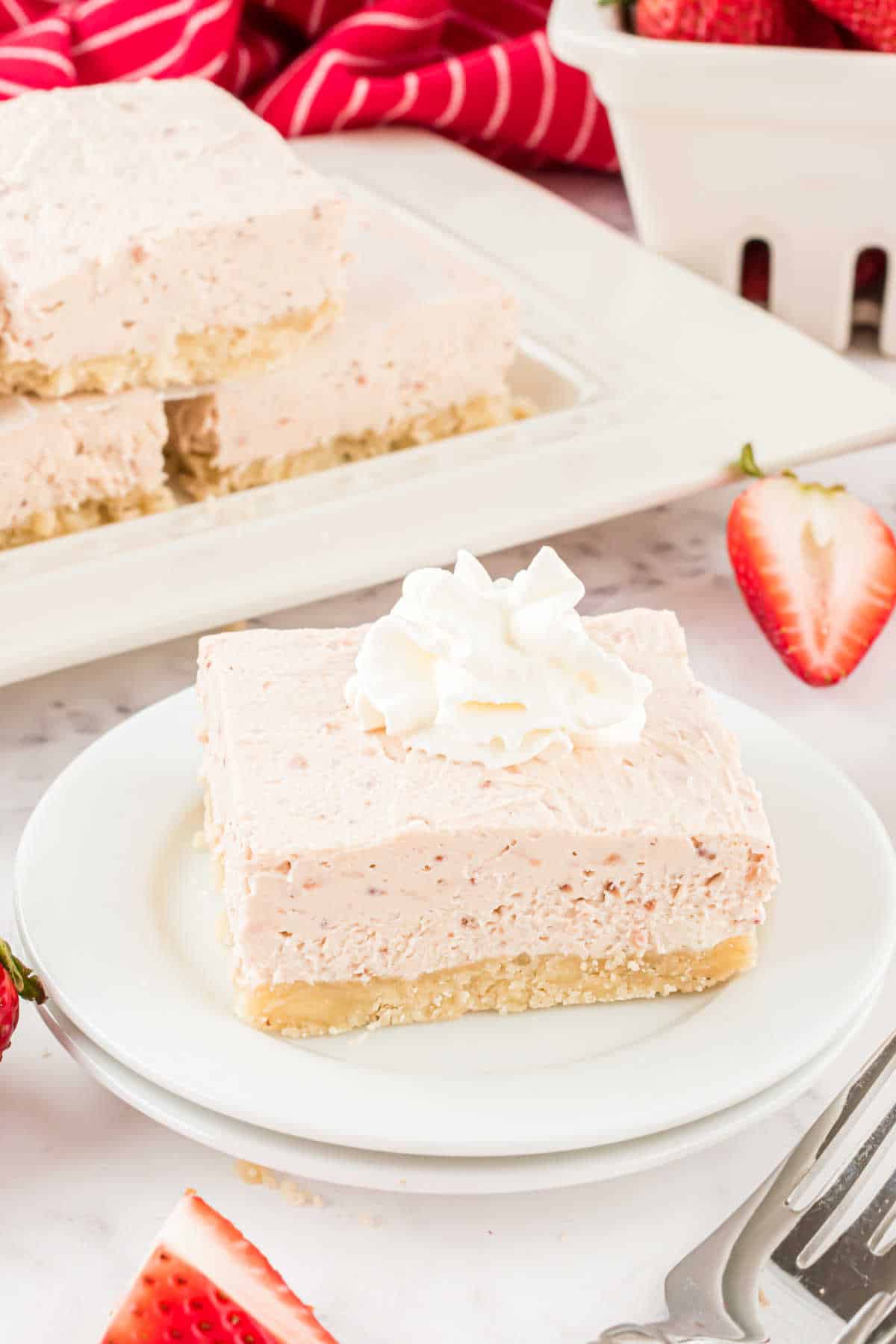 A square slice of strawberry cheesecake topped with whipped cream on a white plate.
