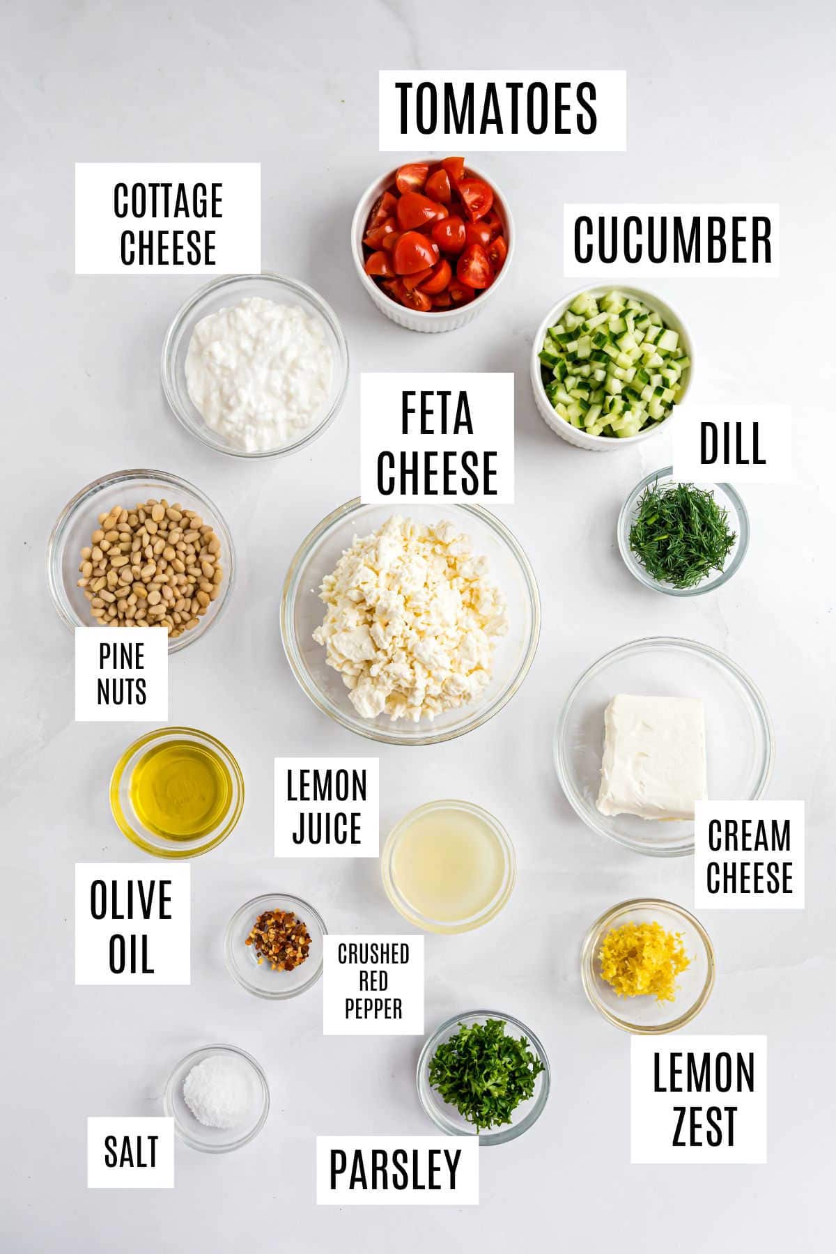 Various bowls with labeled ingredients: tomatoes, cucumber, whipped feta dip, dill, cottage cheese, cream cheese, pine nuts, and more.