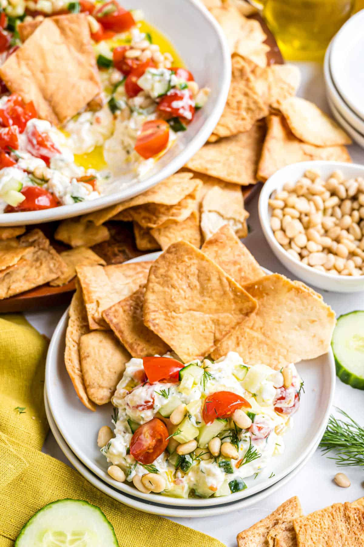 Plate with pita chips and a creamy whipped feta dip topped with tomatoes, cucumbers, herbs, and pine nuts, surrounded by snacks.