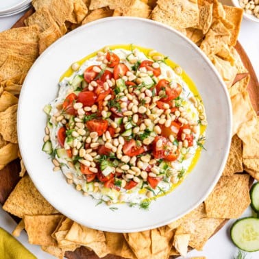 A bowl of whipped feta dip topped with tomatoes, pine nuts, and herbs, surrounded by pita chips.
