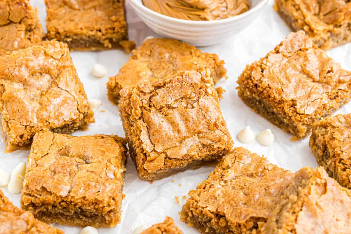 Blondie bars with a crackly top, scattered on parchment paper near white chocolate chips and a bowl of peanut butter.