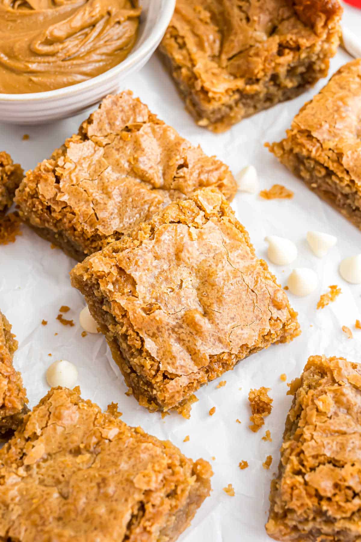 Golden brown peanut butter blondies on parchment with white chocolate chips and a bowl of peanut butter.
