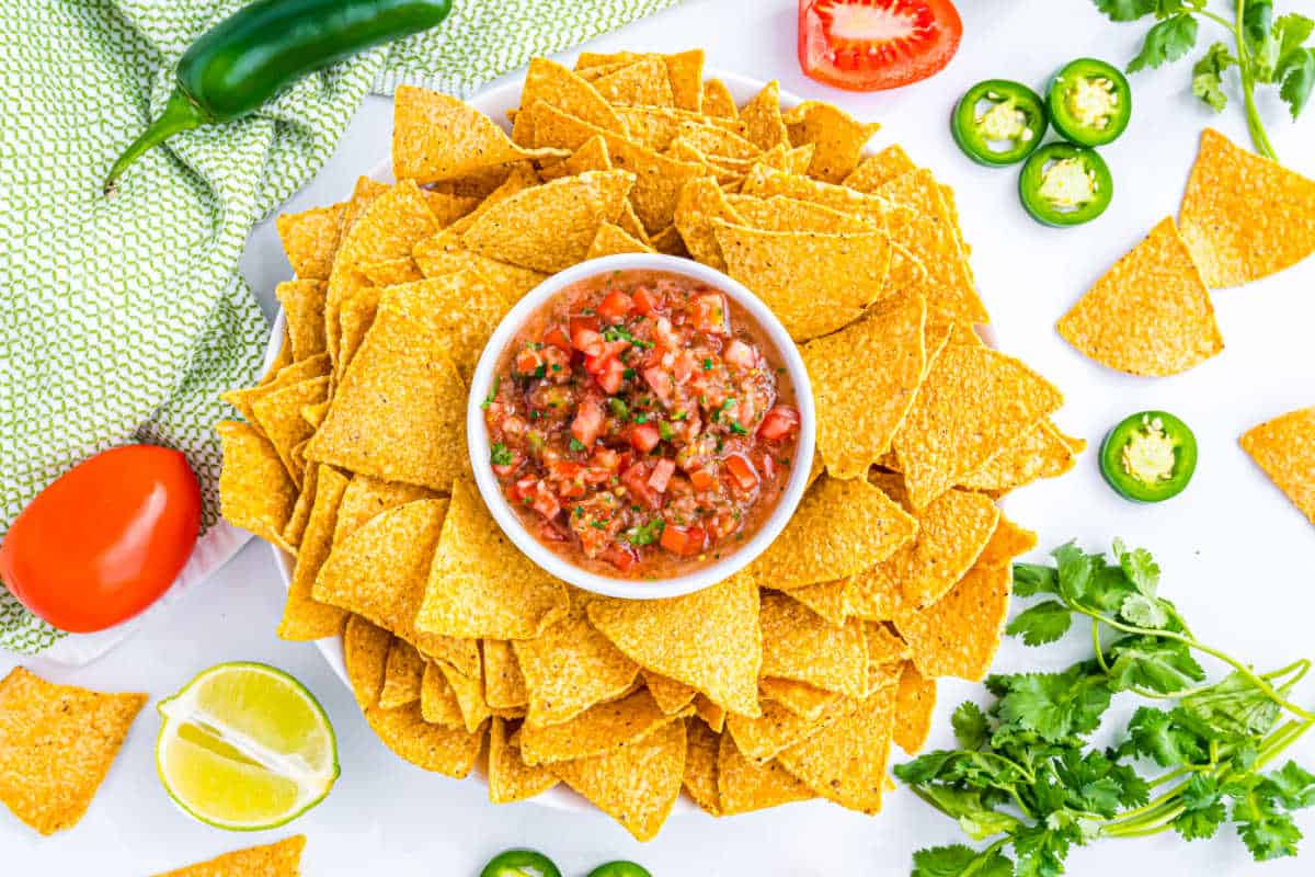 Bowl of salsa in the center of tortilla chips, surrounded by tomatoes, lime, jalapeños, and cilantro.