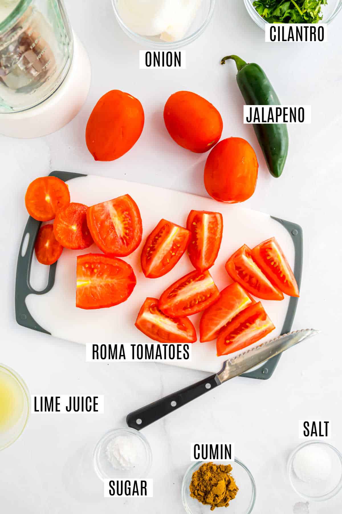 Chopped roma tomatoes on a cutting board, surrounded by labeled salsa ingredients: onion, jalapeno, cilantro, and spices.