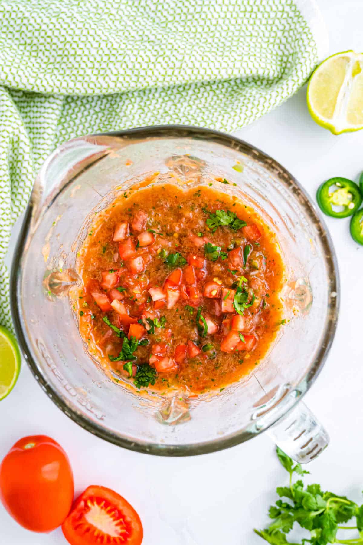 Fresh tomato salsa with cilantro and peppers in a blender, surrounded by tomatoes, lime, and a green towel.