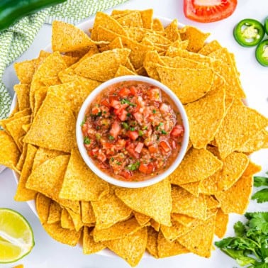 A bowl of salsa surrounded by tortilla chips, with lime, tomato, jalapeño, and cilantro on the side.