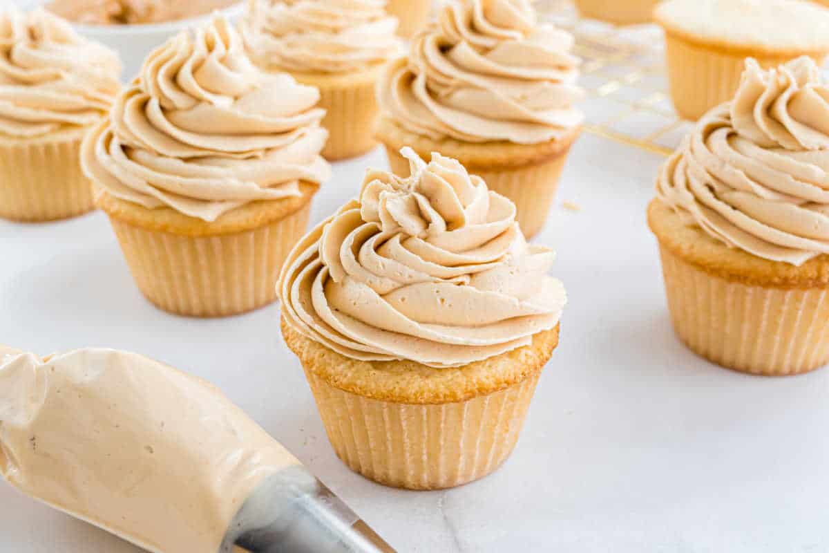 Vanilla cupcakes with swirled light brown frosting on top, with a piping bag in the foreground.