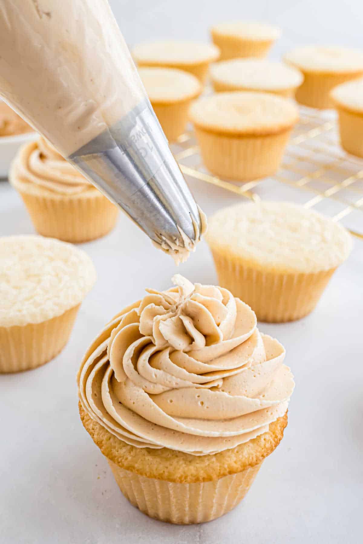Piping creamy frosting onto a vanilla cupcake with unfrosted cupcakes in the background.
