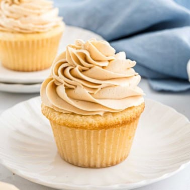 Vanilla cupcake with swirled buttercream frosting on a white plate, with another cupcake in the background.