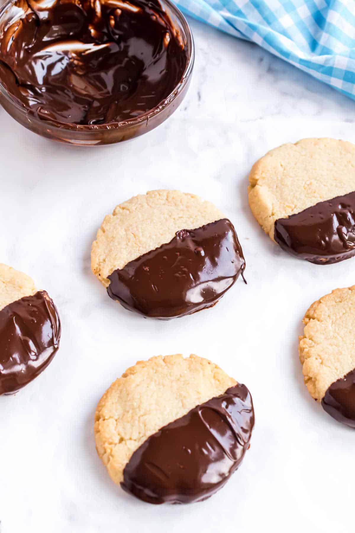 Five cookies dipped halfway in chocolate, with a bowl of melted chocolate nearby on a marble surface.