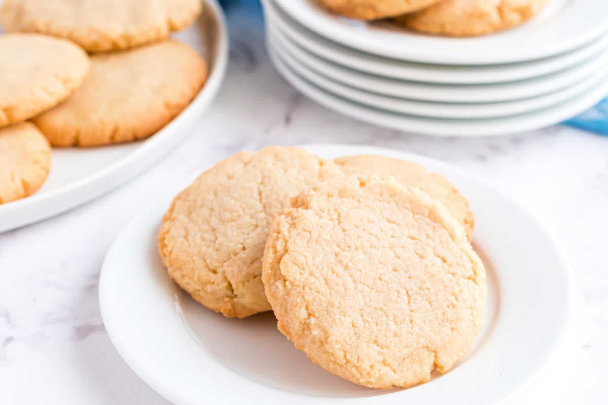 Two sugar cookies on a white plate with more cookies on plates in the background.