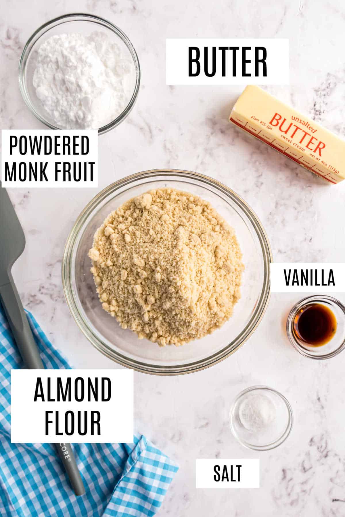Top view of almond flour, butter, powdered monk fruit, vanilla, and salt in bowls on a white surface with a spatula.