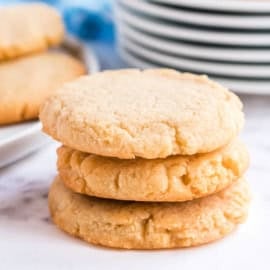 Three sugar cookies stacked on a white surface with a plate of more cookies and stacked dishes in the background.