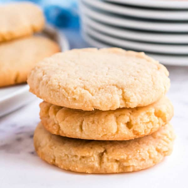 Three sugar cookies stacked on a white surface with a plate of more cookies and stacked dishes in the background.