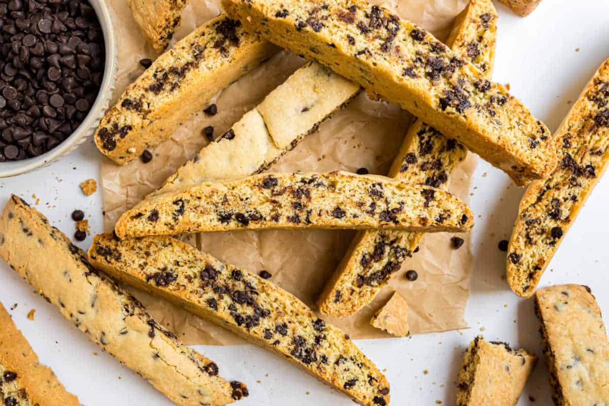 Chocolate chip biscotti scattered on parchment paper with a bowl of chocolate chips nearby.