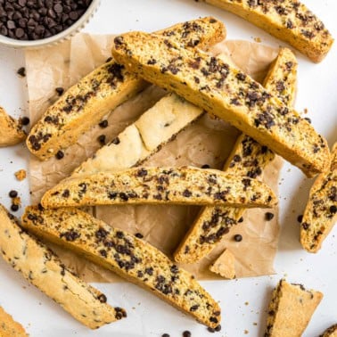 Chocolate chip biscotti scattered on parchment paper, with chocolate chips and a bowl in the background.