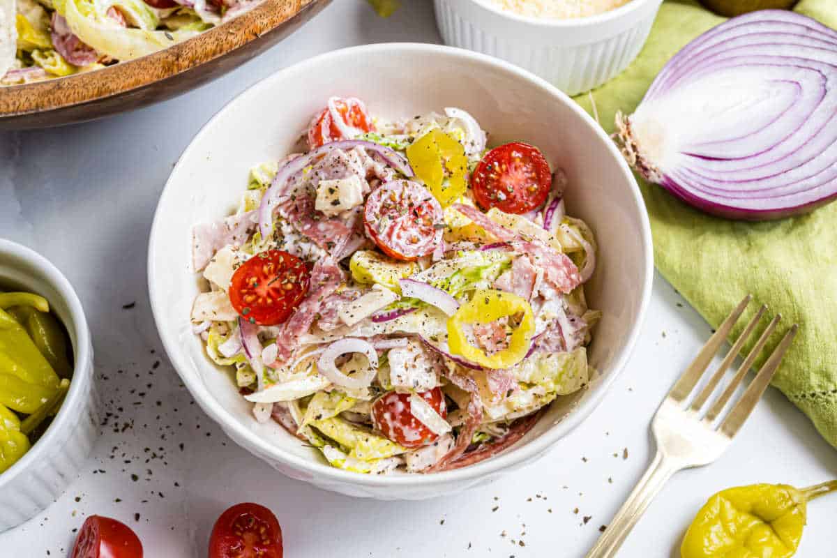 Bowl of Italian chopped salad with lettuce, tomatoes, onions, pepperoncini, and grated cheese on a white table.