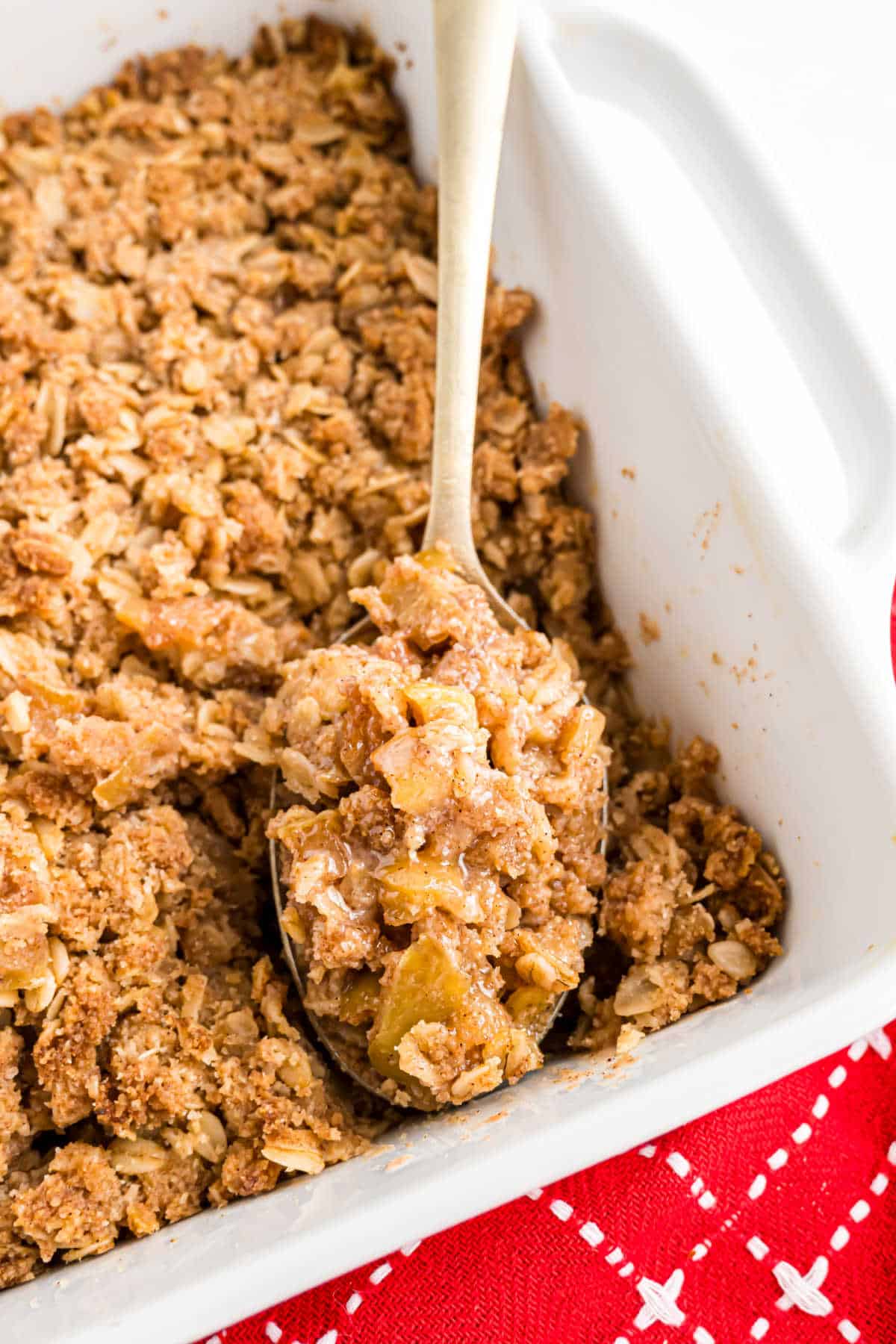 A spoon scooping apple crisp with oat topping from a white baking dish, on a red cloth.