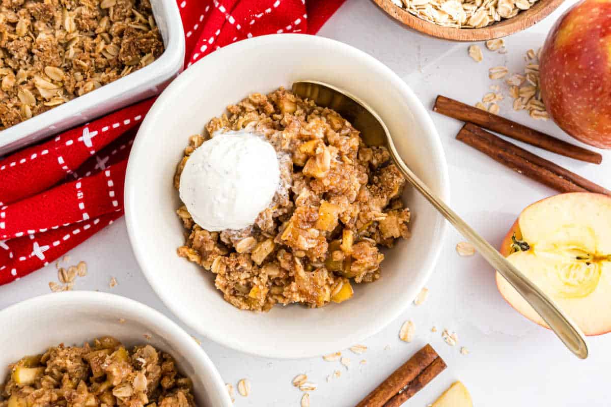 A bowl of apple crisp topped with vanilla ice cream, surrounded by apple slices, oats, and cinnamon sticks.