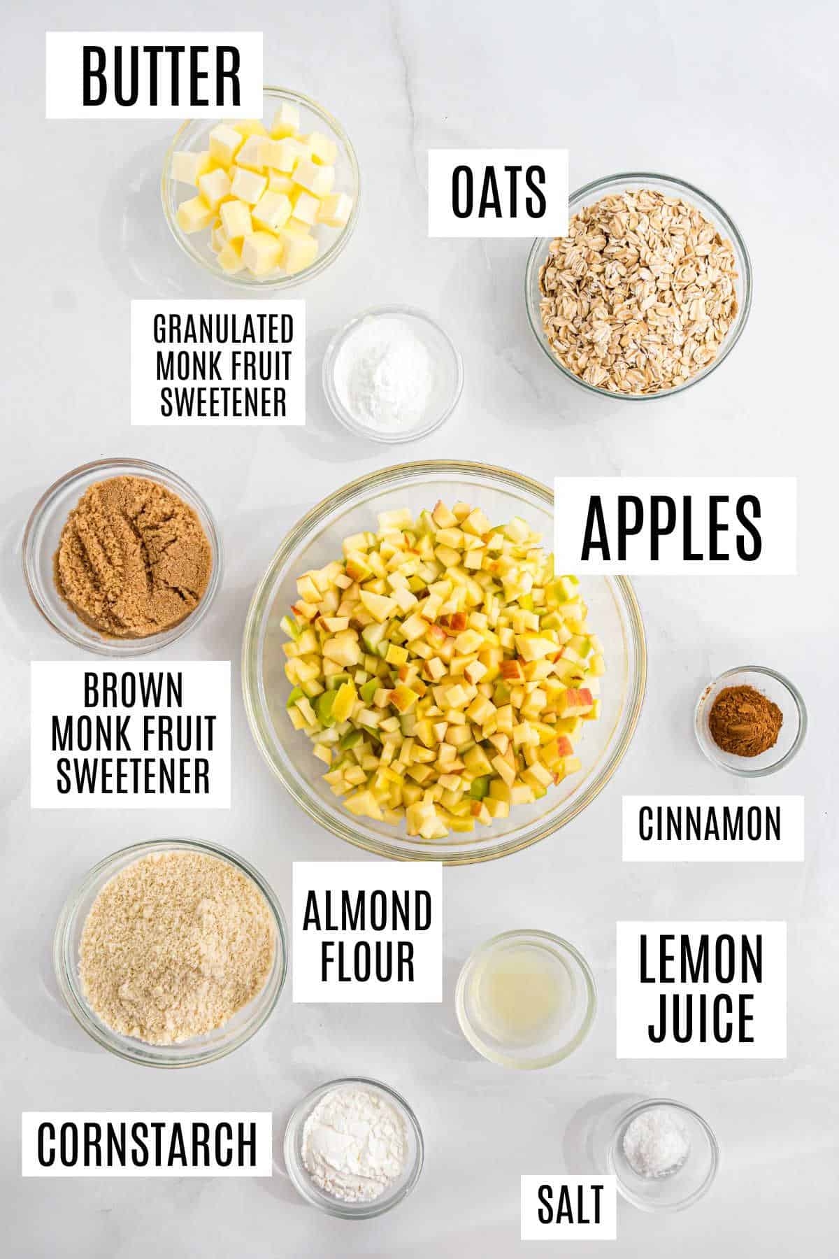 Top view of labeled bowls with ingredients for apple crisp, including apples, oats, butter, sweeteners, and spices.