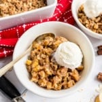 Bowl of apple crisp topped with a scoop of vanilla ice cream, with a spoon and red napkin nearby.