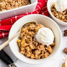 Bowl of apple crisp topped with a scoop of vanilla ice cream, with a spoon and red napkin nearby.