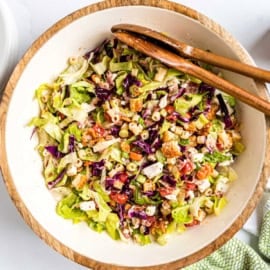 A bowl of chopped salad with pasta, lettuce, tomatoes, croutons, and purple cabbage, with wooden tongs.